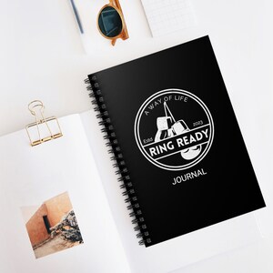 May include: Black spiral-bound journal with the text "Ring Ready Journal" in white. The journal features a circular logo with boxing gloves and the words "A Way of Life" and "2023". The journal is open on a white surface.