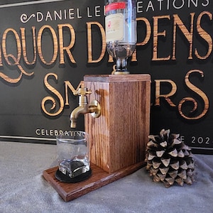 May include: A wooden liquor dispenser with a brass faucet and a clear glass bottle. The dispenser is sitting on a wooden base with a black coaster. The dispenser is in front of a black sign with gold lettering that says "Liquor Dispensers".