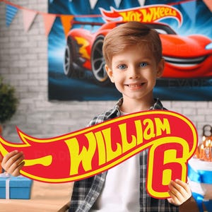 May include: A young boy smiles, holding a red and yellow sign with the text "WILLIAM 6" in a Hot Wheels design. A Hot Wheels car poster is in the background. The scene is set for a birthday celebration, with a cake and gifts visible.
