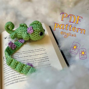 以下が含まれることがあります： 紫色の花が付いた緑色の編みぐるみカメレオンのブックマーク。ブックマークは、「PDF pattern english」と表紙に書かれた本の上に置かれています。