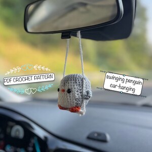 Könnte beinhalten: Ein gehäkelter Pinguin-Anhänger für das Auto mit grau-weißem Körper und orangenen Füßen. Der Pinguin hängt am Rückspiegel eines Autos. Der Text "PDF CROCHET PATTERN" und "swinging penguin car-hanging" ist auf dem Bild zu sehen.