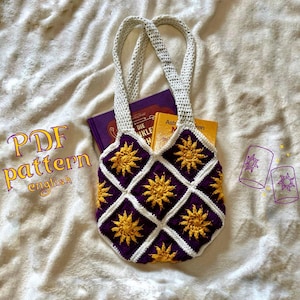 Könnte beinhalten: Eine gehäkelte Tragetasche mit einem violetten und weißen Rautenmuster und gelben Sonnenmotiven. Die Tasche liegt auf einer weißen, flauschigen Oberfläche mit zwei kleinen Bildern einer Sonne und einer Laterne.