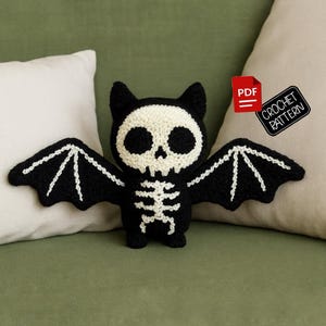 Könnte beinhalten: Eine schwarz-weiße Häkel-Fledermaus mit Totenkopfgesicht und Skelett-Details. Die Fledermaus hat große Flügel und liegt auf einer grünen Oberfläche. Ein rotes PDF-Symbol und ein Etikett "CROCHET PATTERN" sind ebenfalls zu sehen.