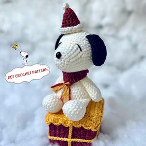 Könnte beinhalten: Gehäkelte Snoopy-Figur mit roter und weißer Weihnachtsmütze und Schal, sitzend auf einer gelben und kastanienbraunen Geschenkbox. Die Figur hat schwarze Ohren und eine schwarze Nase. Die Worte "PDF CROCHET PATTERN" sind in einer weißen Blase.