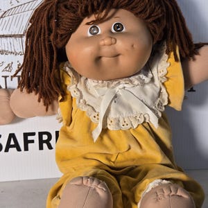 May include: A soft-sculpture doll with brown yarn hair, brown eyes, and a dimpled face. The doll wears a yellow velvet jumpsuit featuring a white lace-trimmed collar with a small white ribbon bow at the center. The doll has light skin.