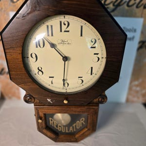 May include: An octagonal wooden wall clock with a dark brown finish and a cream-colored face featuring black Arabic numerals. The clock face displays the text "Warsch Fall River Mass". A lower glass panel contains the word "REGULATOR" in gold.