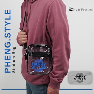 May include: A clear stadium bag with a black zipper and black strap, featuring the blue text HARRY STYLES printed on the front. The bag is worn over a maroon hooded sweatshirt. Additional text in the image includes PHENG.STYLE and Heat Pressed.