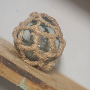 Puede incluir: Un flotador de vidrio redondo envuelto en una red de cuerda trenzada natural. El flotador es de color azul claro con una apariencia blanca nublada. La cuerda es de color marrón claro.