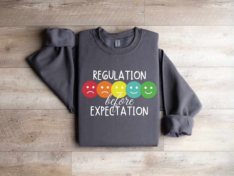 以下が含まれることがあります： 白い文字で「Regulation before Expectation」と書かれた濃いグレーのスウェットシャツ。テキストの上に、カラフルなスマイリーフェイスが5つ表示されています。