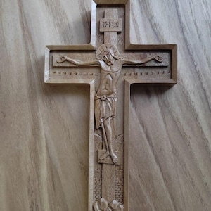 Könnte beinhalten: Ein geschnitztes Holzkreuz, das Jesus Christus darstellt. Das Kreuz ist aus hellbraunem Holz gefertigt und zeigt detaillierte Schnitzereien von Jesus am Kreuz, mit der Inschrift "INRI" oben. Der Hintergrund ist eine Holzoberfläche.