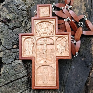 Könnte beinhalten: Ein detaillierter Holzanhänger in Kreuzform mit aufwendigen Schnitzereien, die religiöse Szenen darstellen. Das Kreuz hat eine warme braune Farbe mit einem dunkleren braunen Rand. Eine Holzkette mit Perlen ist am Kreuz befestigt.