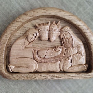 Könnte beinhalten: Eine geschnitzte Holz-Weihnachtskrippe, die Maria, Josef und das Jesuskind in einer Krippe mit Ochse und Esel darstellt. Die Figuren sind aus hellbraunem Holz und in einem gebogenen Rahmen angeordnet.