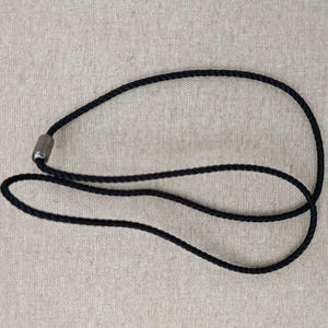 Puede incluir: Un collar de cuerda negra con un cierre metálico. El collar está hecho de un cordón negro tejido apretado. El cierre es una pequeña pieza metálica cilíndrica. El collar se muestra sobre un fondo neutro y texturizado.