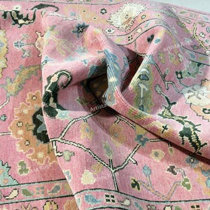 May include: A close-up of a pink rug with an intricate floral pattern. The rug features a variety of colors, including light blue, green, and beige, creating a vintage aesthetic. The rug is folded, showcasing its texture and design.