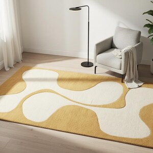 May include: A rectangular area rug with a mustard yellow and cream abstract design. The rug is placed on a light wood floor, with a gray armchair, a black floor lamp, and a plant in the background.