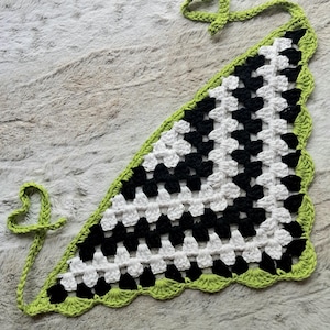 May include: A black and white crocheted triangle scarf with a green border and fringe. The scarf has a checkered pattern and is tied with a green cord.