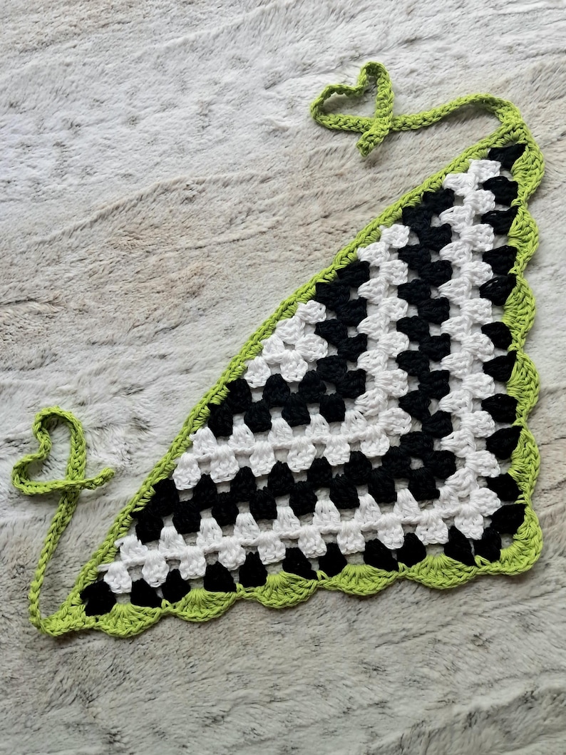 May include: A black and white crocheted triangle scarf with a green border and fringe. The scarf has a checkered pattern and is tied with a green cord.