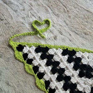 May include: A black and white checkered crochet triangle with a lime green border. The crochet is tied into a heart shape.