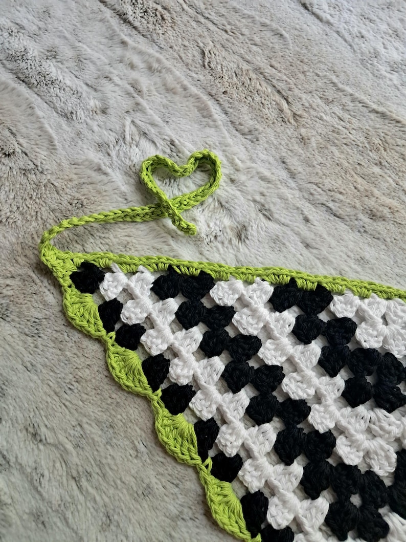 May include: A black and white checkered crochet triangle with a lime green border. The crochet is tied into a heart shape.