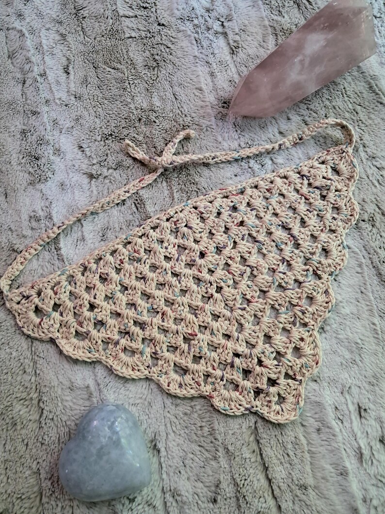 May include: A crocheted beige triangle scarf with multicolored yarn accents. The scarf has a scalloped edge and is tied with a bow.