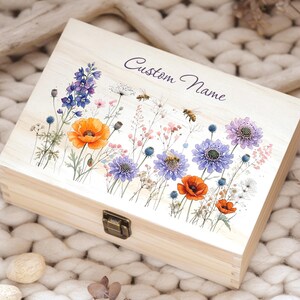 May include: A light-colored wooden box with a floral design featuring orange poppies, purple and blue flowers, and bees. The words "Custom Name" are written on the lid. The box has a metal clasp.