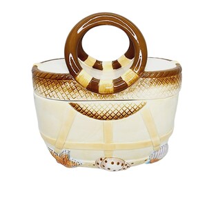 May include: A ceramic cookie jar shaped like a beach bag with a brown rope handle, white and brown stripes, and a brown net design. The jar is decorated with seashells and starfish.