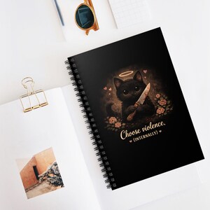 May include: Black spiral-bound notebook with a cartoon black cat illustration holding a knife, and a halo. The text "Choose violence. (internally)" is printed below the cat. The notebook is on a white surface.