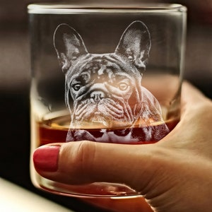 Puede incluir: Un vaso de vidrio transparente con un Bulldog Franc&eacute;s grabado en el lateral. El vaso est&aacute; lleno de l&iacute;quido &aacute;mbar y est&aacute; siendo sostenido por una mano.