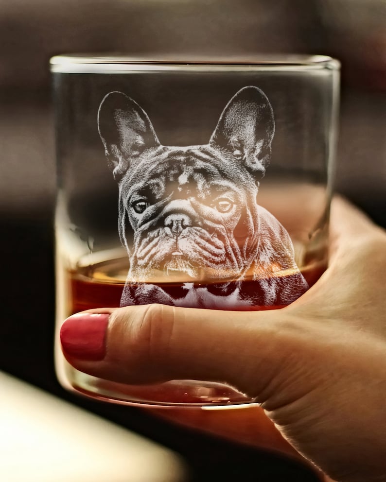 Puede incluir: Un vaso de vidrio transparente con un Bulldog Franc&eacute;s grabado en el lateral. El vaso est&aacute; lleno de l&iacute;quido &aacute;mbar y est&aacute; siendo sostenido por una mano.