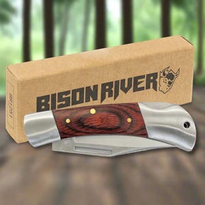 Könnte beinhalten: Ein Klappmesser mit einer Klinge aus Edelstahl und einem Griff aus rotbraunem Holz, verziert mit Messingnieten. Das Messer wird mit seiner Pappschachtel präsentiert, auf der der Schriftzug "BISON RIVER" und ein Logo zu sehen sind.