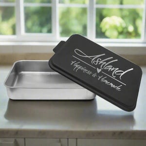 May include: A rectangular baking pan with a black lid. The lid has the word "Ashland" in a script font, with "Happiness is Homemade" below it. The pan is silver and sits on a countertop.