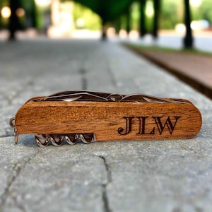 May include: A wooden multi-tool with a corkscrew, knife, and other tools. The wood is a warm brown color, and the initials "JLW" are engraved on the side. The tool is closed and resting on a stone surface.