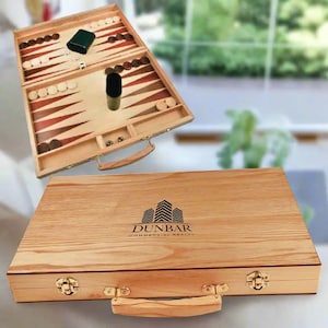 Puede incluir: Un juego de backgammon de madera, abierto para revelar el tablero con fichas marrones y blancas. La caja cerrada presenta el texto "DUNBAR COMMERCIAL REALTY" y un logotipo de edificio. Incluye dados y cubilete.
