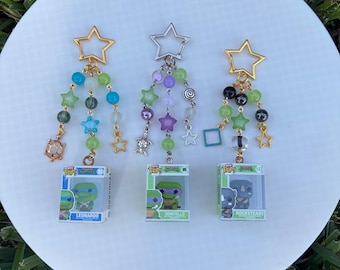 May include: Three keychains with star-shaped tops and colorful beads. Each keychain has a miniature Teenage Mutant Ninja Turtles Funko Pop figure in a clear display case. The figures are Leonardo, Donatello, and Rocksteady.