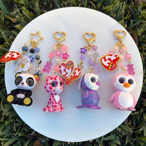May include: A collection of colorful keychains featuring various animal figurines, including a panda, leopard, owl, and penguin. Each keychain has a gold-colored heart or star-shaped clasp and decorative beads. The figurines are set on a white circular surface.