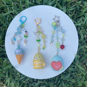May include: Three colorful keychains with charms. The first keychain has a blue and white ice cream cone charm with a cat face. The second keychain has a yellow and white mug charm with a yellow bear face. The third keychain has a blue heart charm with a red heart on it.