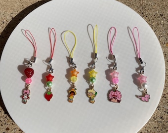 May include: Six colorful phone charms featuring cartoon characters and beads. The charms are pink, red, yellow, green, and blue. Each charm has a small, colorful star bead and a larger bead in a different color. The charms are attached to a thin, pink string.