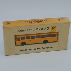 May include: A model of a yellow Deutsche Post AG bus in its original box. The box is tan with black text and the Deutsche Post AG logo. The bus has black wheels and windows. The box reads "Modellautos für Sammler".