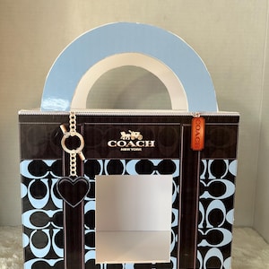 May include: A blue and black Coach gift bag with a white handle and a cut-out window. The bag has a Coach logo and a heart-shaped charm.