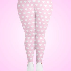 May include: Pink leggings with a repeating pattern of white hearts. The leggings are shown from the knees down, with white shoes visible at the bottom. The background is a gradient of pink.