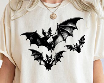 May include: Cream-colored t-shirt featuring a black illustration of several bats in flight. The bats have detailed wings and are arranged in a dynamic composition. The shirt is a casual style, suitable for Halloween or gothic themes.