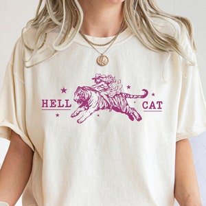 May include: A white t-shirt with a pink graphic of a tiger with a woman riding on its back. The graphic includes the text "HELL CAT" and stars.