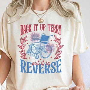 May include: A white t-shirt with a blue and red graphic that says "Back it up Terry put it in reverse". The graphic features a wheelchair with fireworks and a wreath of leaves.