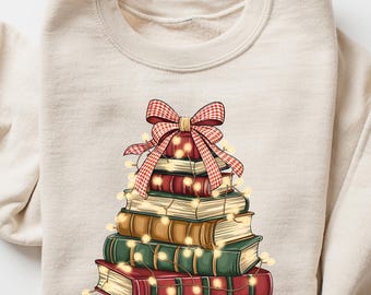 May include: Cream-colored sweatshirt featuring a graphic of a stack of books decorated as a Christmas tree. The books are red, green, and tan, adorned with a red and white plaid bow and string lights. A cozy, festive design.
