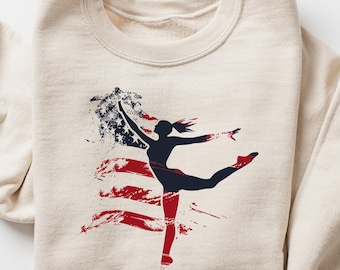 May include: A beige sweatshirt with a graphic print of a silhouette of a dancer in front of an American flag. The flag is red, white, and blue with a distressed look.