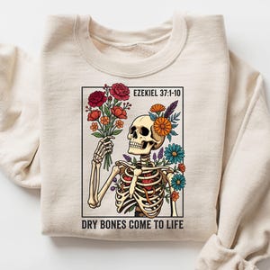 May include: A beige crewneck sweatshirt featuring a graphic print of a skeleton holding a bouquet of colorful flowers. The text EZEKIEL 37:1-10 appears at the top, and the text DRY BONES COME TO LIFE is printed at the bottom of the rectangular frame.