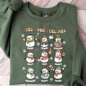 May include: Military green sweatshirt with the text "FEEL YOUR FEELINGS" and illustrations of snowmen expressing different emotions. The bottom of the sweatshirt reads "MILITARY GREEN".