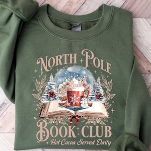 May include: Military green sweatshirt with the text "North Pole Book Club" and a graphic of a snow globe, a mug of hot cocoa, and a snowman. The sweatshirt is a crew neck style.