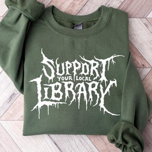 May include: An olive green crewneck sweatshirt featuring white graphic text on the front that reads SUPPORT YOUR LOCAL LIBRARY in a stylized, jagged, dripping font. The garment has long sleeves with ribbed cuffs and a ribbed neckline.