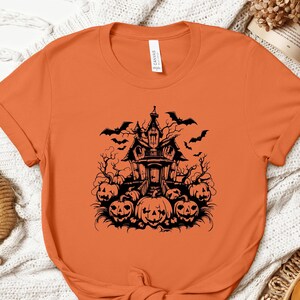 May include: Orange t-shirt featuring a black Halloween design. The graphic includes a haunted house, bats, and jack-o'-lanterns. The shirt is made of a soft material and is perfect for the spooky season.